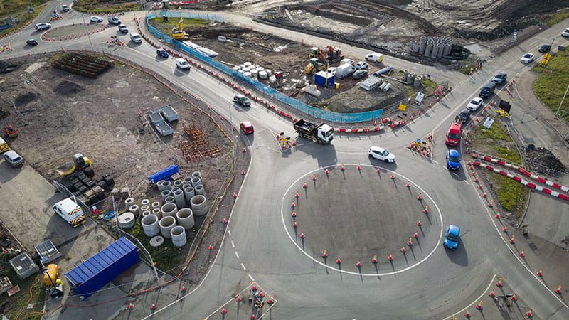 An aerial view of a roundabout construction site with the roundabout in the centre. Multiple vehicles are navigating the roundabout and surrounding roads. The site is fenced off with cones and barriers.