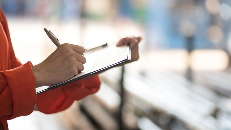 A person holding a clipboard and writing on it with a pen. The background is blurred.