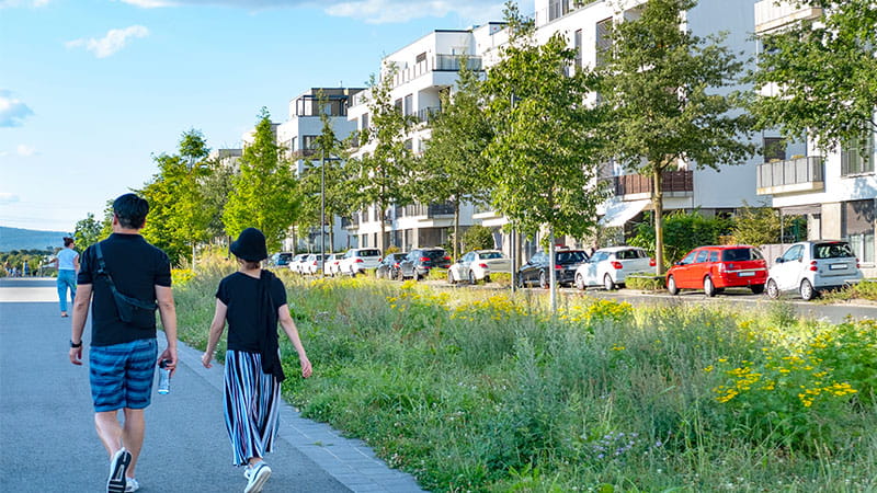 A man and a woman walking through a sustainable housing development with modern white apartment buildings, green space and trees separating the roads, and parked cars in the background.