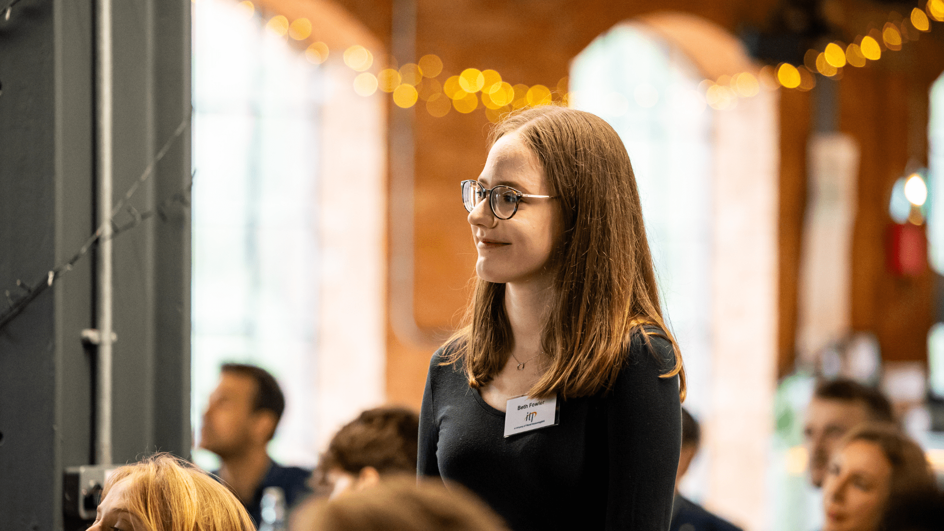 A person with shoulder-length hair stands among a seated audience in a warmly lit indoor venue with exposed brick walls and tall arched windows. The person is wearing glasses, a dark long-sleeved top, and a visible name badge. Soft string lights are draped across the background, creating a festive atmosphere. Other attendees can be seen seated nearby, and the setting suggests a formal event, presentation, or workshop.