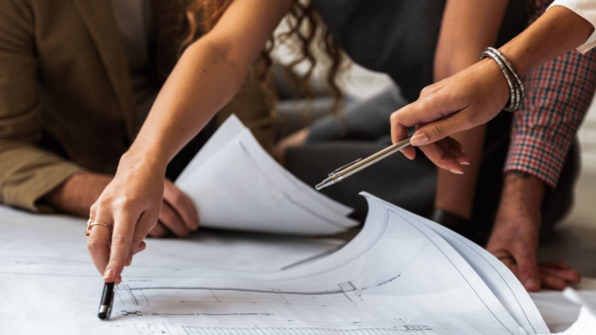 People reviewing technical drawings and plans together, pointing at documents spread across a table during a project planning meeting.