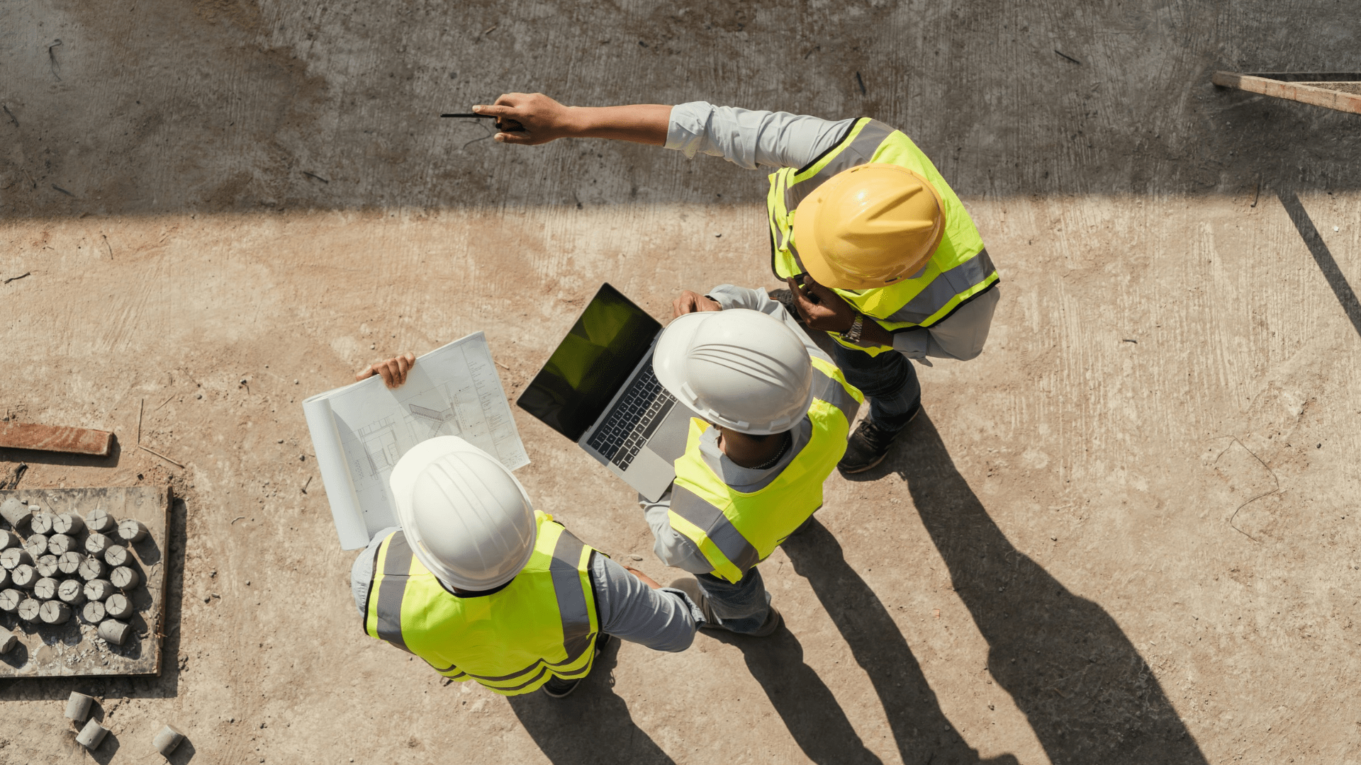 Construction professionals in high-visibility clothing reviewing plans and a laptop while inspecting a construction site from above.