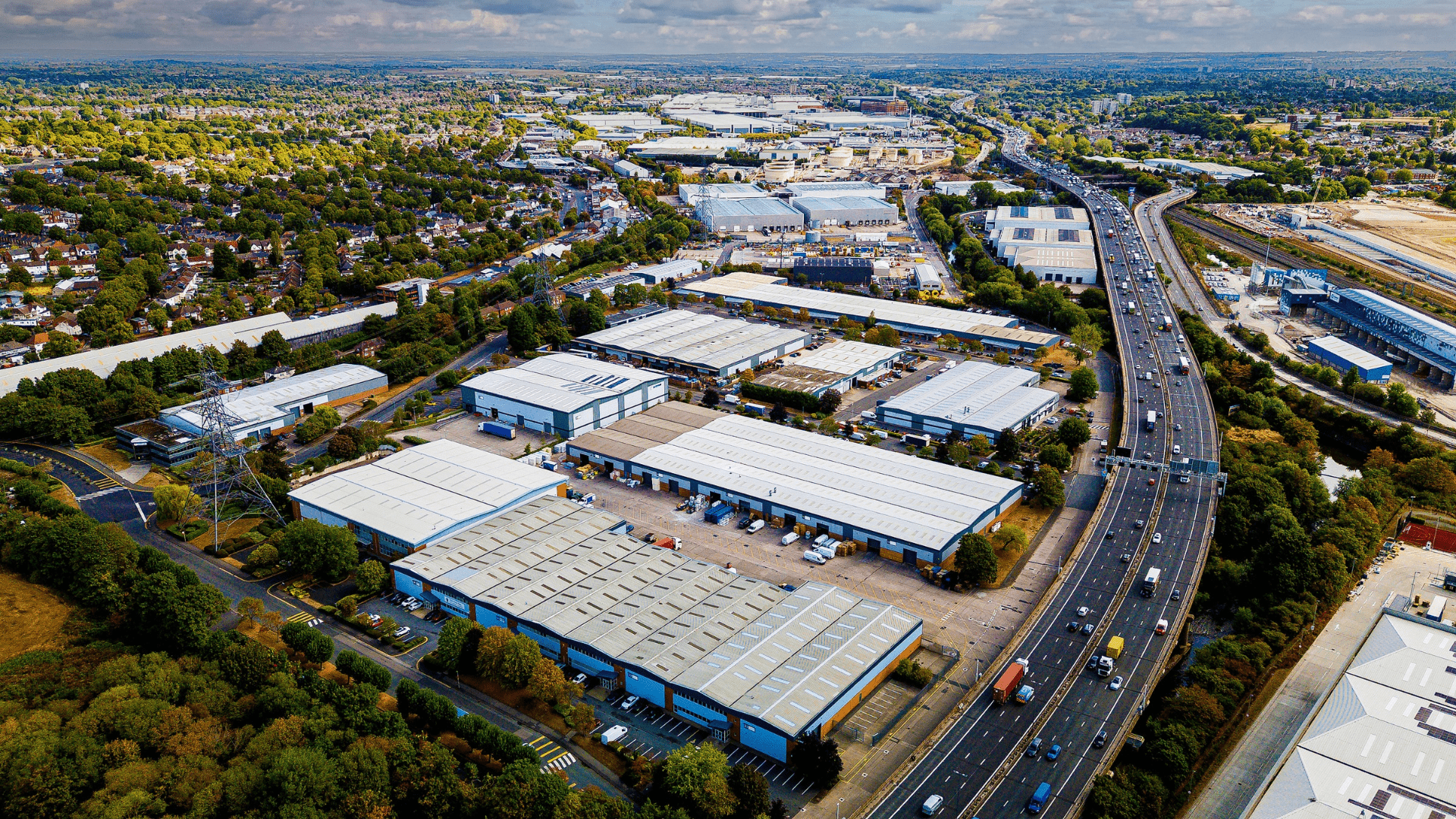 Aerial view of an industrial and logistics park alongside a major motorway, showing warehouses, transport infrastructure and surrounding urban development.