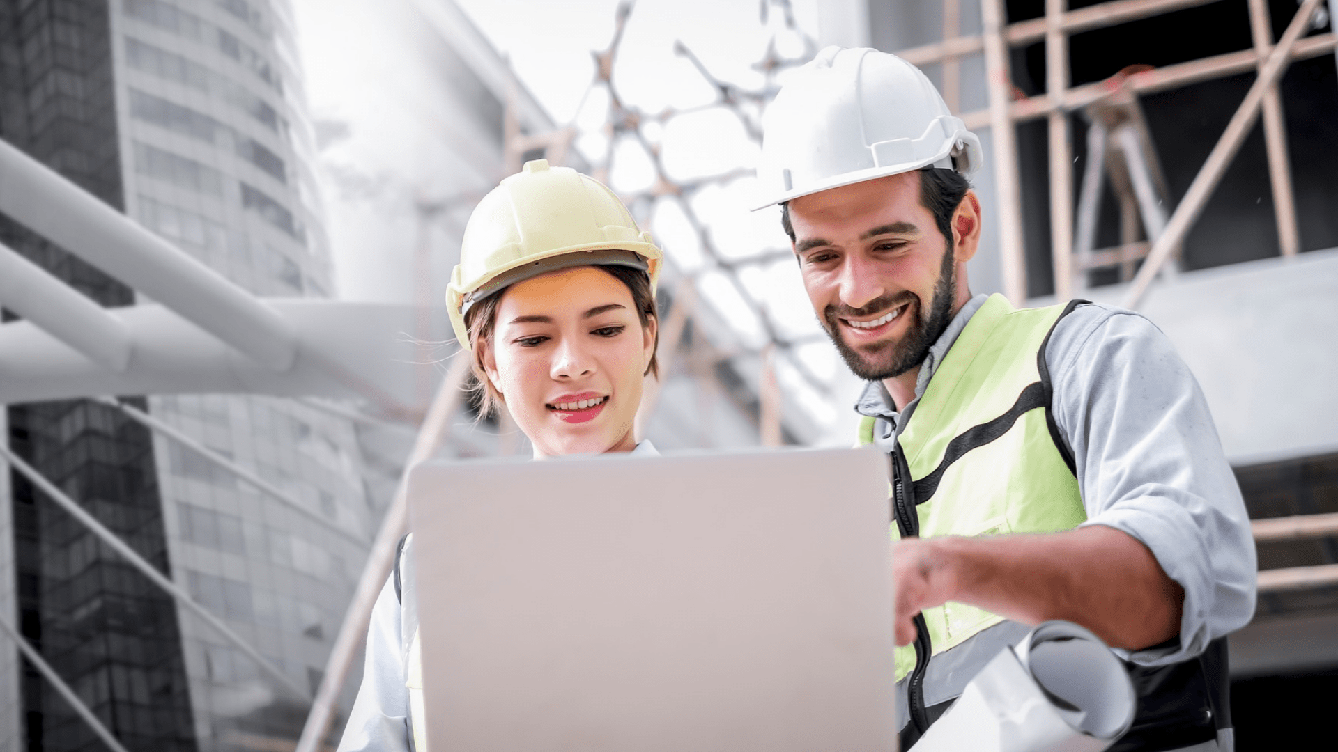 Construction professionals wearing safety helmets and high-visibility clothing reviewing project information on a laptop at an active construction site.