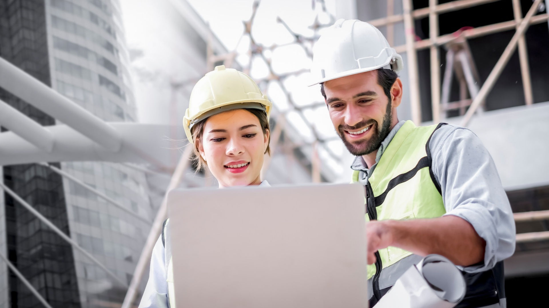 Construction professionals wearing safety helmets and high-visibility clothing reviewing project information on a laptop at an active construction site.