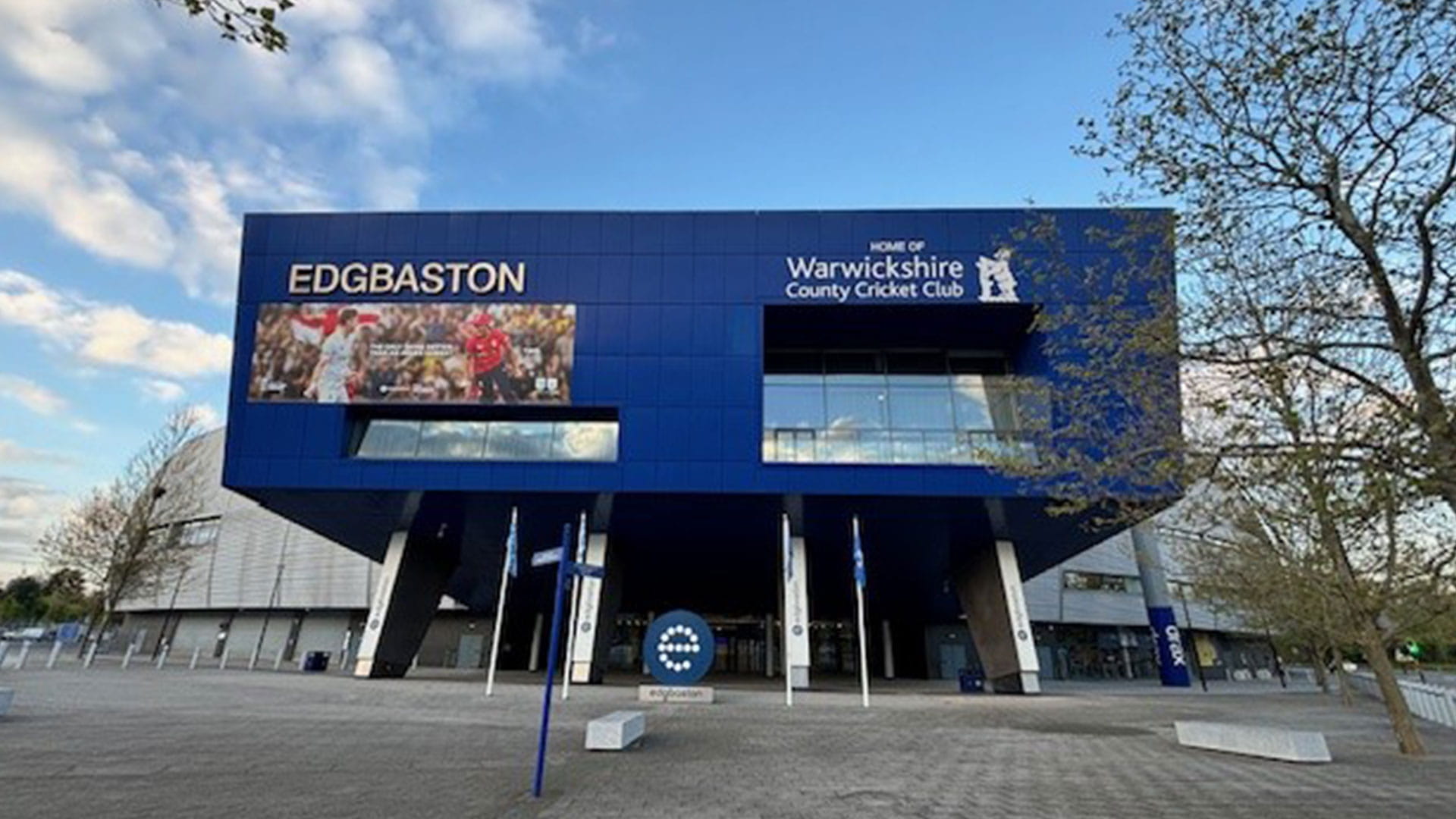 The image shows the exterior of Edgbaston, home of Warwickshire County Cricket Club. The building has a modern design with a large blue facade featuring the name "EDGBASTON" and an image of cricket players in action.