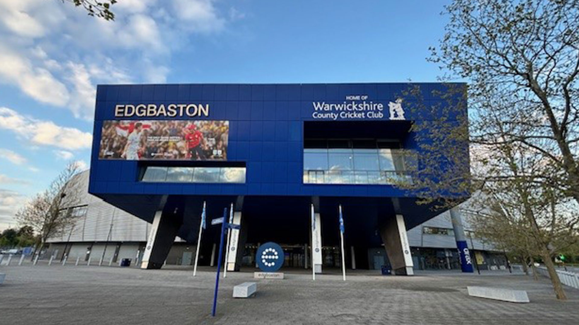 The image shows the exterior of Edgbaston, home of Warwickshire County Cricket Club. The building has a modern design with a large blue facade featuring the name "EDGBASTON" and an image of cricket players in action.